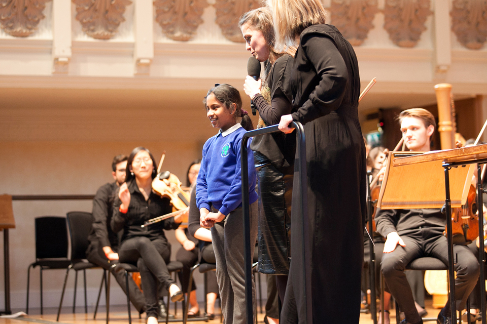 Two female presenters on the stage, standing in a conductors podium, with an Asian girl on stage in a school uniform.